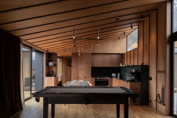 A warm, timber-lined open-plan interior centred on a dark billiards table, with a compact kitchen and dining area beyond. The space features extensive wood wall and ceiling panelling, dark cabinetry, integrated appliances, pendant lighting, tall glazing, and a refined recreational-lounge atmosphere.