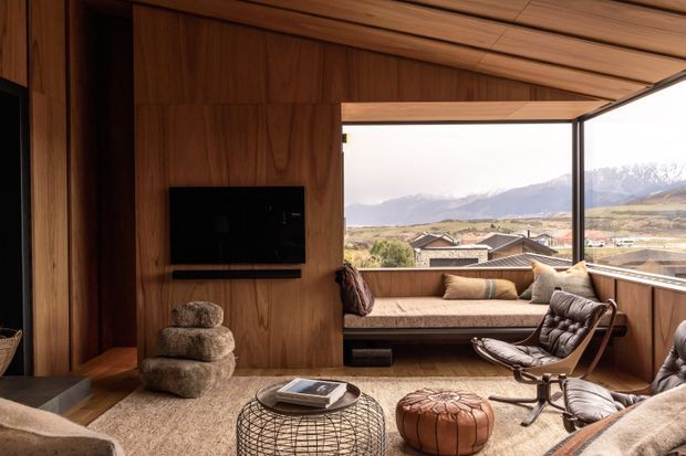 Warm timber-lined living room with a sloping wood ceiling, built-in television wall, low seating, leather lounge chairs, textured rug, round wire coffee table and leather ottoman. Full-height corner glazing opens the room to a broad mountain and rural valley view, creating a sheltered lodge-like interior.