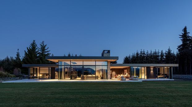 A wide dusk exterior view of a low-slung, single-storey contemporary residence set against a dense forest. The home has a strongly horizontal composition, extensive floor-to-ceiling glazing, dark roof planes, warm interior lighting, and several sheltered outdoor living areas opening onto a broad lawn.