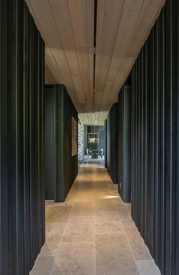 A long, narrow interior hallway with strong linear perspective. Dark vertical timber wall panelling and deep door recesses frame a warm timber-panelled ceiling, while large rectangular beige stone or porcelain floor tiles lead toward a distant seating area. The space has a dramatic, refined, materially rich contemporary character with subdued lighting.