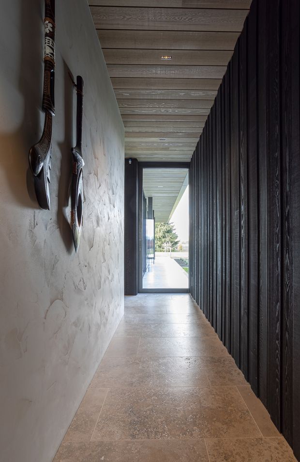 A narrow, atmospheric entry corridor leading to a glazed exterior door. The hallway has a textured plaster wall with elongated decorative wall sculptures, a dark vertically ribbed timber wall on the right, a pale timber-lined ceiling with exposed linear boards, and large-format warm stone-look floor tiles. Daylight enters from the doorway at the end, creating a strong contrast between the dark interior and bright exterior.