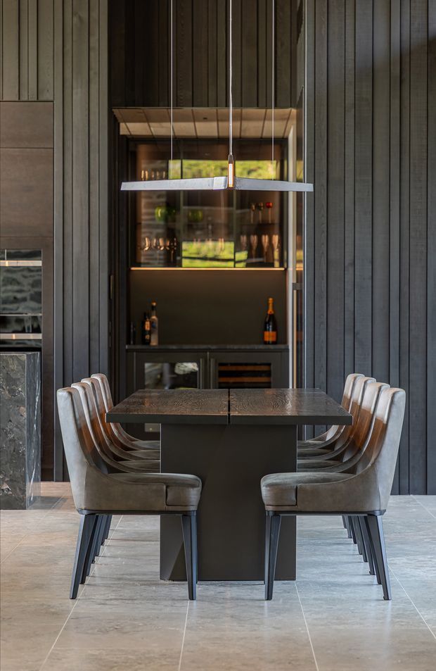 A sophisticated dining area with a long dark rectangular dining table and upholstered dining chairs arranged on both sides. The room features dark vertical timber wall panelling, a built-in illuminated bar or drinks cabinet behind the table, suspended linear pendant lights, a stone or porcelain tiled floor, and a partial fireplace or stone feature at the left edge. The composition is symmetrical, refined, and moody with contemporary detailing.