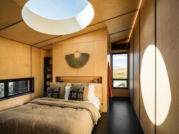 Compact, warmly lit bedroom clad extensively in natural timber plywood panels, with a built-in bed, dark bedding, integrated shelving, narrow black-framed windows, and a large circular skylight or ceiling opening. A narrow passage leads to a rear window with a view of the surrounding landscape. The room has a refined, cabin-like contemporary design with strong built-in joinery and dramatic daylight.