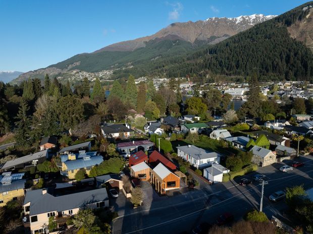 Aerial view over a suburban residential neighbourhood with detached homes, mature trees, streets and rooftops in the foreground. A broad forested mountain range with patches of snow rises prominently behind the town, creating a scenic alpine setting under clear blue skies.