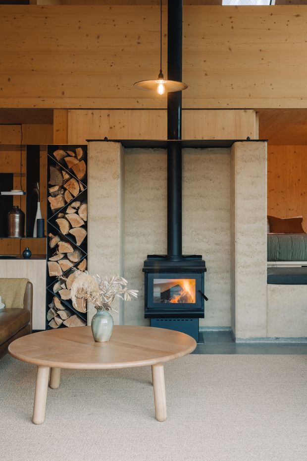 A warm, rustic living room centred on a freestanding black wood-burning stove with a tall flue, set against a pale textured masonry hearth and timber-lined walls. A built-in firewood storage niche, low seating, round light-wood coffee table, pendant light, and soft neutral carpet create a cosy cabin-like interior.