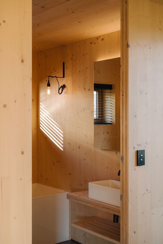 A compact, warm timber-lined bathroom viewed through an open doorway. The room features a white freestanding bathtub, a white vessel basin on an open timber vanity with a slatted lower shelf, a small window with horizontal blinds, and a minimalist black wall-mounted light fixture. Sunlight creates striped shadows across the timber wall.