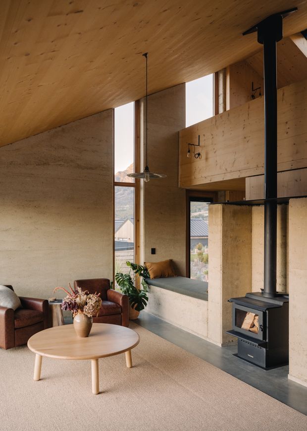Warm contemporary living room with extensive exposed timber lining to the sloped ceiling, textured earthen or concrete-like walls, tall vertical glazing, a black freestanding wood-burning stove, built-in window seat, leather lounge chairs, and a round timber coffee table. The room has a cozy, natural material palette and views toward a residential landscape.