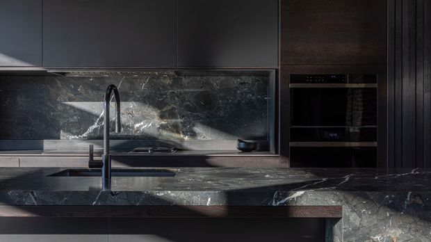 A dramatic, contemporary kitchen featuring dark flat-panel cabinetry, a large island with a deep veined dark stone benchtop, matching stone splashback, integrated oven and microwave, and a tall black gooseneck tap over the sink. The palette is predominantly charcoal, black, and deep brown with striking pale stone veining and restrained metallic accents.