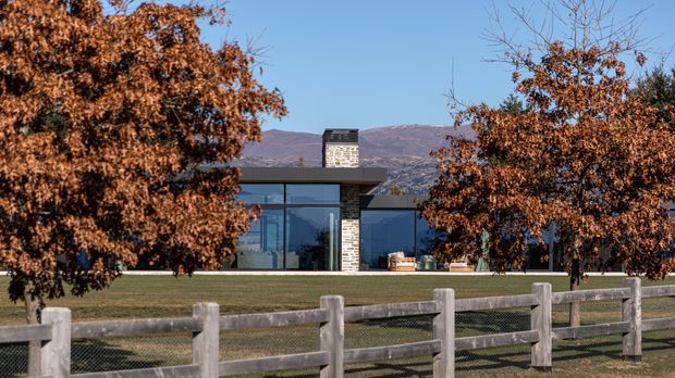 A low, modern single-storey house set in a landscaped rural or semi-rural setting, viewed from across a timber post-and-rail fence. The building has extensive dark-framed floor-to-ceiling glazing, a flat roof, and a prominent rectangular stone-clad chimney or masonry feature. Autumnal trees frame the facade, with dry grass and mountain or hill slopes visible behind it.