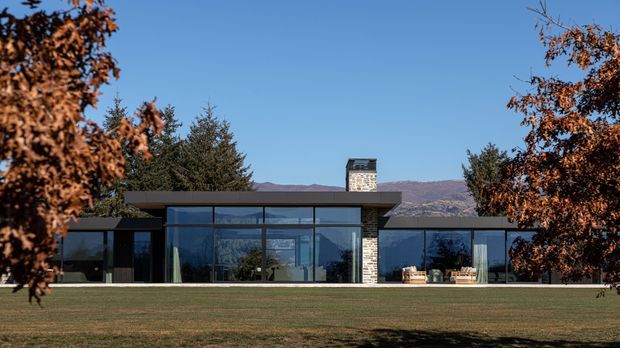 A low, single-storey contemporary house set in a broad landscaped lawn, with extensive floor-to-ceiling glazing, dark horizontal roof forms, and a central stone-clad chimney or vertical feature. Mature trees frame the facade, while a mountain ridge is visible beyond the property.