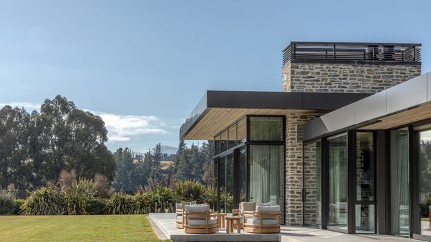 A contemporary multi-level residential exterior with a projecting flat-roofed wing, extensive black-framed glazing, warm timber soffits, and a prominent exposed stone-clad tower. An outdoor seating area sits on a paved terrace overlooking landscaped grounds and mature trees.
