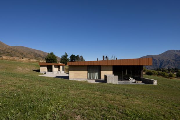 A low-profile contemporary single-storey house set in an open grassy valley, with a long horizontal form, flat roof, dark glazing, pale masonry walls, and a prominent rust-brown metal roof fascia or cladding band. Detached outbuilding elements sit to the left, while dry hills and mountains form the distant backdrop under a clear blue sky.