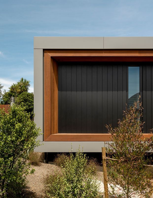 Close-up exterior view of a contemporary building facade, featuring dark vertical cladding framed by warm timber reveals and a broad light-grey metal or rendered surround. A narrow vertical window reflects the surrounding landscape. The building is slightly elevated above a landscaped gravel garden, with timber fencing and dense planting visible around it.