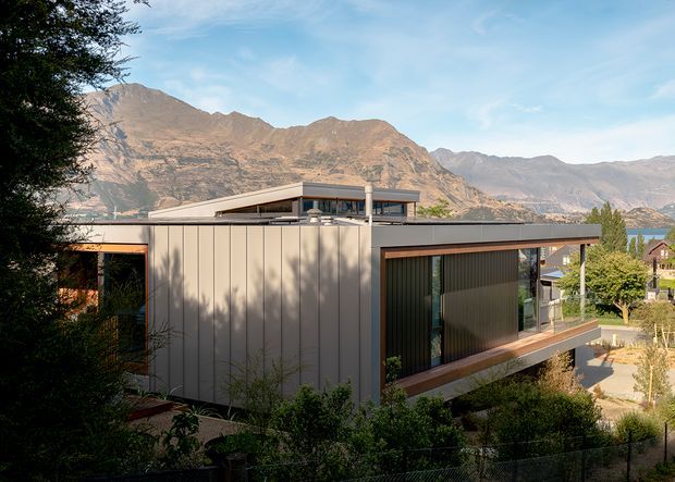 Exterior view of a contemporary, flat-roofed multi-level house set against a rugged mountain landscape. The building features clean rectilinear volumes, extensive glazing, dark horizontal roof and balcony bands, vertical light-coloured facade panels, and landscaped outdoor areas around the home.