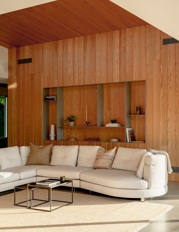 A warm, refined living room featuring a large cream sectional sofa, nested metal coffee tables, and a full-height built-in timber wall with integrated shelving and concealed storage. The wall and ceiling are clad in richly coloured vertical-grain wood, creating a cohesive architectural feature. Sunlight enters from the side, casting long shadows across the pale rug and floor.