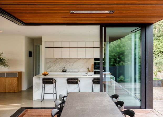 A refined open-plan kitchen and dining area with a large white island, three bar stools, marble-look splashback, and minimalist handleless cabinetry. The dining table occupies the foreground, while full-height glazed sliding doors open toward a leafy exterior. A warm timber-lined ceiling contrasts with the white joinery, pale flooring, and dark aluminium framing.
