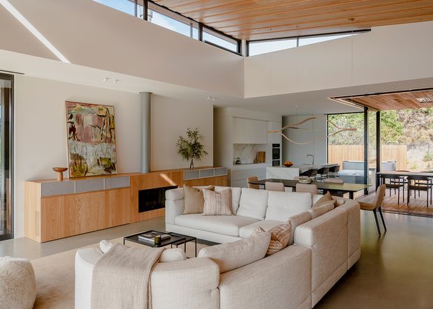 A bright, spacious open-plan living area with a large cream sectional sofa, low black coffee table, and timber media/storage console. The living room opens directly to a modern white kitchen and dining area, with full-height glazing leading to an outdoor deck. Architectural features include a high ceiling, clerestory glazing, warm timber ceiling lining, polished concrete flooring, and restrained contemporary joinery.