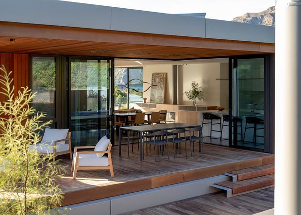 A contemporary indoor-outdoor entertaining area with a covered timber-lined ceiling, generous dark-framed sliding glass doors, and a timber deck furnished with a long dining table and chairs. The deck connects directly to an open-plan interior containing a kitchen or bar area and additional dining furniture. A mountain or rocky hillside is faintly visible beyond the building.