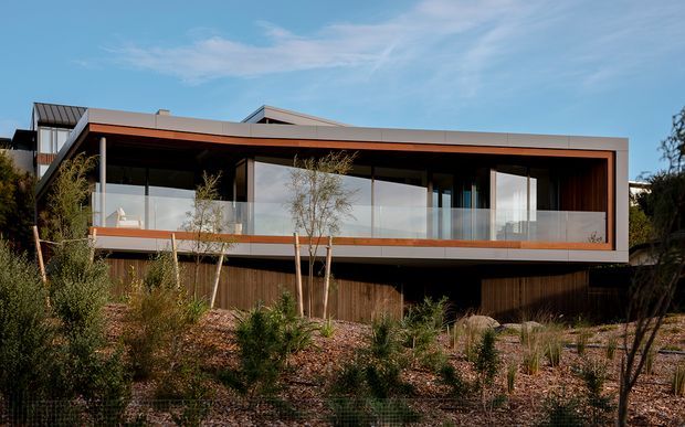 A contemporary two-level residential facade with a long cantilevered upper volume, dark timber cladding, warm orange-brown trim, and extensive floor-to-ceiling glazing. A glass-balustraded terrace runs across the front, while the lower level is partly screened by vertical timber elements. The house is set within a landscaped, dry-climate garden under a bright blue sky.