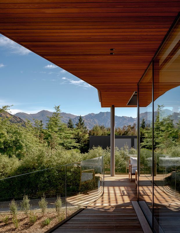 A modern elevated timber deck or balcony terrace beneath a broad cantilevered timber-lined roof soffit. Full-height glazed walls reflect the deck and surrounding vegetation, while a transparent glass balustrade frames expansive mountain views. A small outdoor seating area is visible, with lush planting and a dry landscaped edge around the deck.