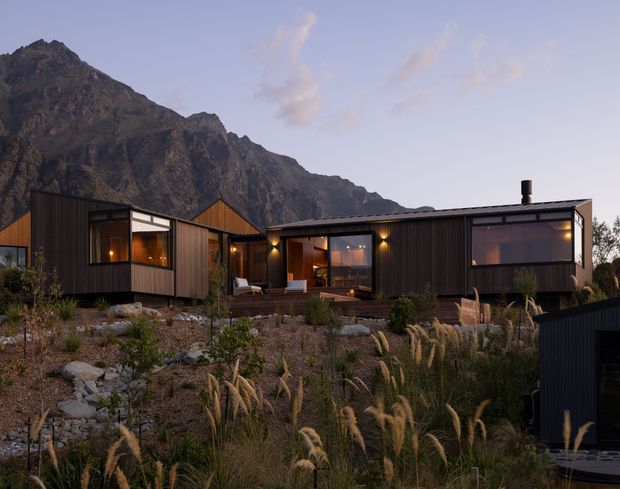 A low, modern multi-volume house set against a dramatic mountain backdrop. The dark vertical-clad exterior has large glazed openings, warm interior lighting, a covered central outdoor connection, and a landscaped rocky garden with ornamental grasses in the foreground.