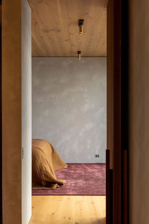 A warm, minimalist bedroom viewed through an open doorway. The room has a pale textured plaster wall, a timber-lined ceiling with small black-and-brass downlights, warm timber flooring, and a muted rose-purple area rug. A low bed or mattress is partially visible at the left, covered with a mustard-gold quilt. Dark timber door framing contrasts with the light interior.