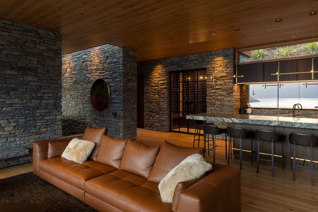 A warm, sophisticated open-plan living room and home bar with a large caramel leather sectional sofa in the foreground. Rough stacked-stone walls and a timber-lined ceiling create a richly textured interior, while a stone-topped bar with dark cabinetry, stools, pendant lighting, and a glass-front wine storage area occupies the rear. The space has a moody, luxurious lodge-like character.