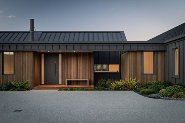 Single-storey contemporary residential facade viewed from the driveway, featuring dark standing-seam metal roofs and cladding, warm vertical timber wall boards, black-framed windows and entry door, a covered recessed entrance, and restrained low-maintenance landscaping with gravel and planting beds.