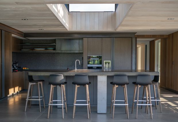 A spacious contemporary kitchen centered on a long dark island with seating for five or more. The room features floor-to-ceiling dark timber cabinetry, a textured dark backsplash, integrated appliances, a pale stone or concrete-like island face, and a timber-lined ceiling with a large skylight. Minimal black bar stools and warm natural light create a refined, modern atmosphere.