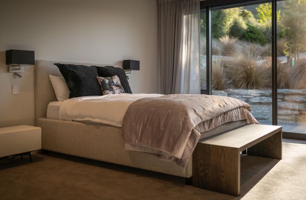 A warmly styled contemporary bedroom with a low upholstered bed, layered neutral bedding, dark textured cushions, wall-mounted bedside lights, and a simple timber bench at the foot of the bed. Full-height glazing and curtains open onto a landscaped garden, while a soft carpet and muted beige palette create a calm, refined atmosphere.