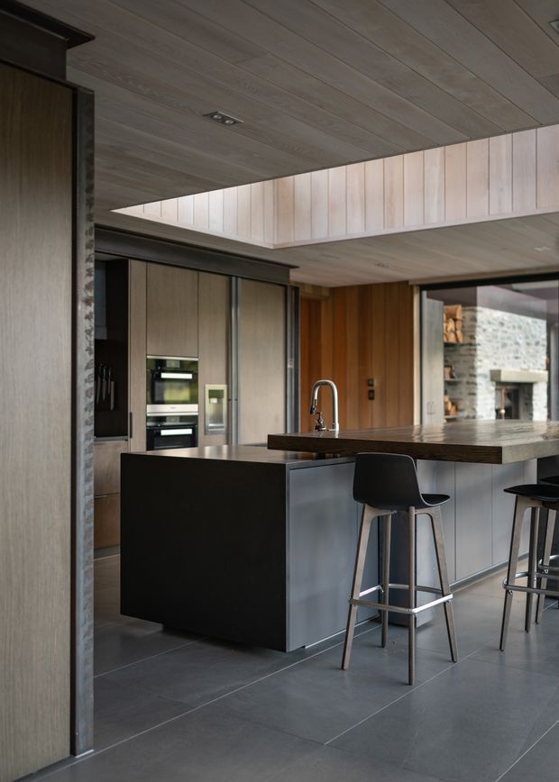 A contemporary kitchen with a large dark island, integrated appliances, timber-toned cabinetry, a deep wood benchtop, and bar seating. The room features extensive timber wall and ceiling lining, dark grey tiled flooring, and a broad clerestory-style opening that brings in natural light. A stone fireplace or masonry feature is visible through the opening at right.