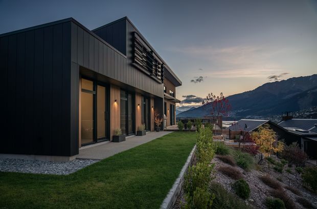 Contemporary dark-clad multi-level house on a sloping landscaped site, photographed at dusk. The angular facade features vertical metal cladding, tall glazed doors, projecting volumes, and horizontal sun-shading screens. A lawn and planted garden terrace overlook neighbouring rooftops and a dramatic mountain backdrop.