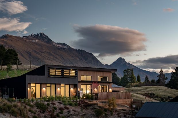 A contemporary two-storey dark-clad house set on a sloping alpine site, photographed at dusk. The home features large warm-lit windows, timber-toned entry and balcony elements, landscaped terraces, and dramatic rugged mountains in the background.