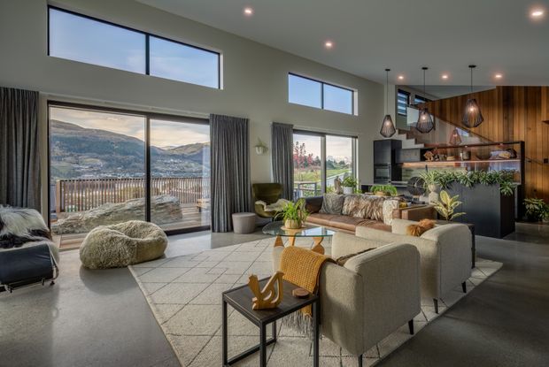 A spacious, contemporary open-plan living room with a lounge and bar area. Floor-to-ceiling sliding glazing opens to a balcony with mountain views, while high clerestory windows bring in natural light. The room features a polished concrete floor, neutral upholstered seating, a large rug, timber wall panelling, open shelving, and sculptural pendant lights over the built-in bar.