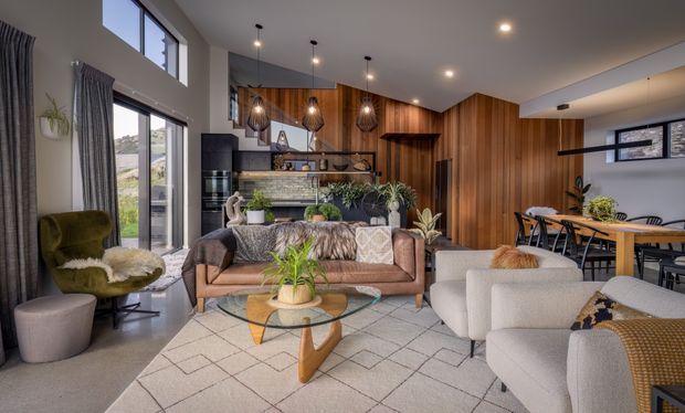 A spacious open-plan living area with a mid-century-inspired seating arrangement, large corner windows, a timber-lined kitchen and dining zone, pendant lighting, indoor plants, and a soft neutral rug. The room has a warm, layered, contemporary feel with high ceilings and abundant natural light.