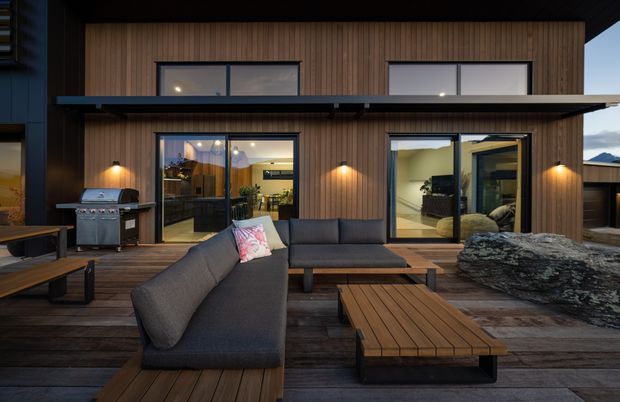 Contemporary two-storey home facade viewed from a timber outdoor deck at dusk. The exterior features warm vertical timber cladding, dark metal framing and roof elements, large glazed openings, and an outdoor entertaining area with built-in barbecue, sectional lounge seating, and timber tables. Interior rooms are visible through the glazing, with a mountainous landscape in the distance.