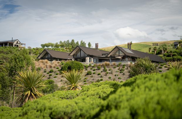 Wide exterior view of a low-profile residential house set within an extensively landscaped, dry-climate garden. The home has multiple dark pitched gable roofs, exposed timber-like structural framing, large glazed openings, and dark exterior finishes. Layered shrubs, grasses, yucca-like plants, and rolling open hills surround the property.