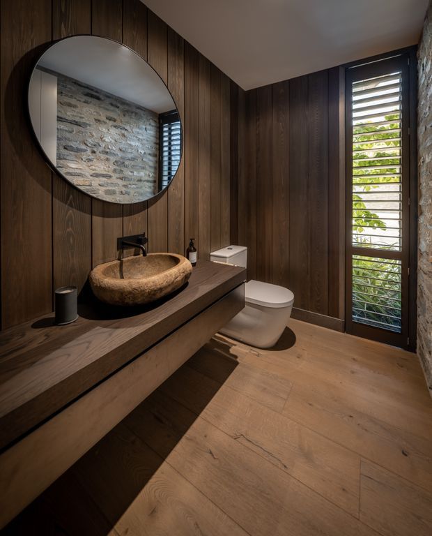 A compact, moody bathroom with dark vertical timber wall panelling, matching timber vanity and flooring, a sculptural natural stone vessel basin, black tapware, a wall-mounted toilet, and a large round mirror. A tall louvred glazed door or window brings in filtered daylight, while exposed rough stonework is visible on the adjacent wall.