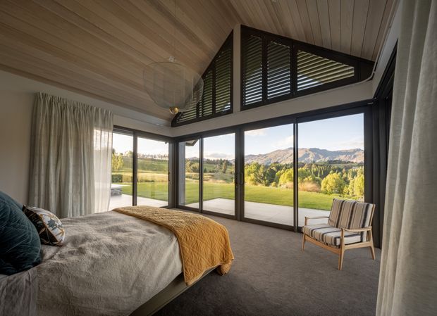 A serene bedroom with a low-profile bed, textured grey bedding, mustard throw, and upholstered armchair. The room features warm timber-lined ceilings, full-height dark-framed glazing, sheer curtains, and high clerestory windows with timber louvres. A broad rural mountain landscape is visible beyond the glazing.