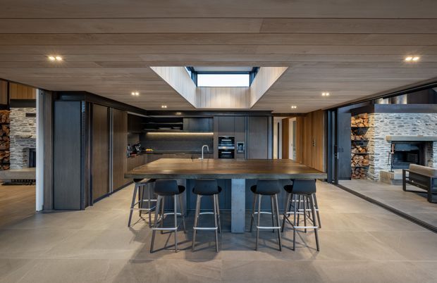 A spacious contemporary kitchen centred on a large dark stone island with timber-toned countertop and seating for four. The kitchen features dark integrated cabinetry, built-in appliances, a linear timber ceiling, recessed downlights, and a dramatic central skylight. Full-height glazing or openings connect to a living area with a substantial stone fireplace and stacked firewood visible on the right. Large-format pale floor tiles create a refined, neutral finish.