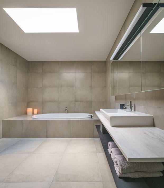 A contemporary minimalist bathroom with large-format taupe-grey tiles covering the walls and floor. The room includes a built-in freestanding-style bathtub, a wide stone vanity with a raised rectangular basin, wall-mounted taps, mirrored cabinetry, recessed ceiling lighting, and open shelving with folded towels.
