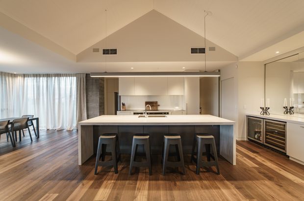 Open-plan contemporary kitchen and dining area with a large central island and four stools, white handleless cabinetry, integrated appliances, timber flooring, and a compact built-in wine refrigerator. The room has a vaulted ceiling, pendant wiring, recessed lighting, expansive curtains, and warm natural light.