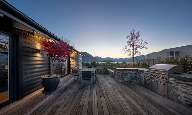 A spacious timber deck beside a dark-clad modern home, featuring outdoor dining furniture, potted trees, stone retaining walls, and a built-in stainless-steel barbecue kitchen. The elevated terrace overlooks a lake and mountainous landscape at dusk.