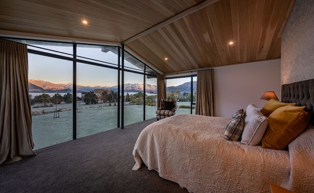 A warm, contemporary bedroom with a large upholstered bed, layered neutral and ochre bedding, dark carpet, timber-lined ceiling, and full-height black-framed glazing. The expansive windows overlook a lake or river valley with mountainous terrain beyond.