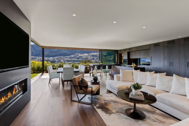 Open-plan contemporary living and dining area with a kitchen at the rear, featuring expansive corner glazing and a panoramic mountain-and-valley view. The room has dark timber built-in cabinetry, wide timber flooring, a large cream sectional sofa, a linear fireplace beneath a wall-mounted television, and a dining table positioned beside the glazing.