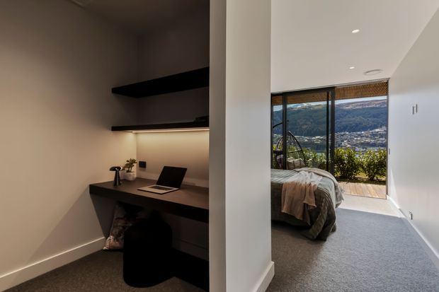 A contemporary interior showing a compact built-in home office nook on the left, with dark desk cabinetry, floating shelves, integrated task lighting and a laptop. Beyond the dividing wall is a carpeted bedroom with a bed and full-height glazed doors opening to a balcony or exterior view. The space has white walls, recessed ceiling lights, dark joinery and mountain scenery visible through the glazing.