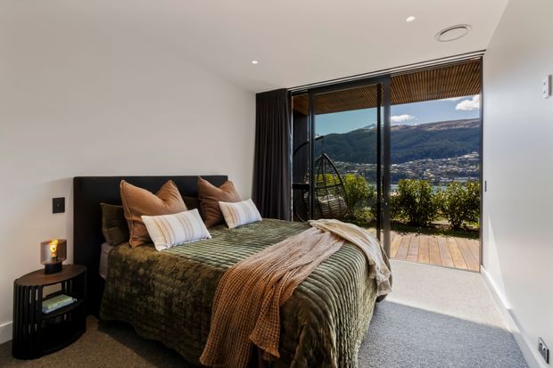 A contemporary bedroom with a low-profile bed dressed in layered olive-green and warm brown bedding, positioned beside a small black side table. Full-height glazed sliding doors open to a timber deck and a mountain-and-valley view, with dark curtains, a simple white ceiling, recessed lighting, and grey carpet. A hanging egg chair is visible outside.