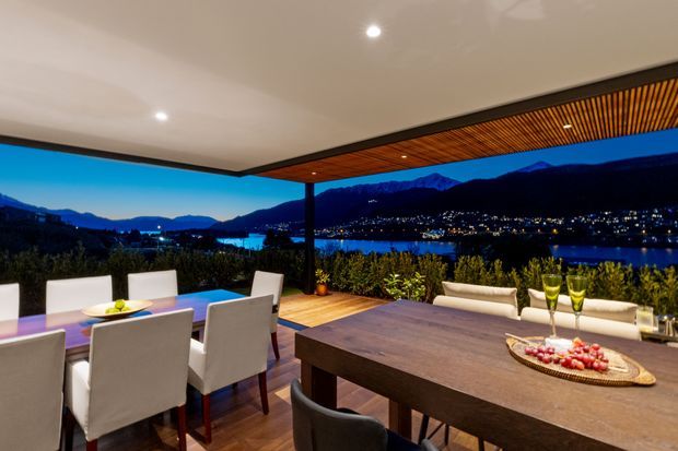 A covered outdoor dining and entertaining terrace at dusk, with a large timber dining table, upholstered dining chairs, a secondary dining setting, and built-in bench seating. The terrace has a flat white ceiling, a timber slatted soffit overhang, dark structural posts, timber decking, and open views across a lake toward illuminated mountains.