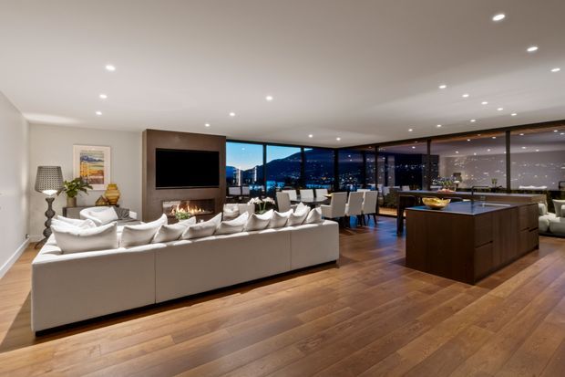 Spacious open-plan contemporary living area with a large cream sectional sofa, dark timber feature media wall and fireplace, dining area, and a substantial dark wood kitchen island. Full-height corner glazing overlooks a mountainous landscape at dusk, while recessed ceiling lighting and warm timber flooring create a polished, inviting atmosphere.
