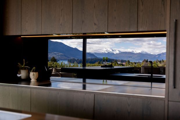 A dark, refined contemporary kitchen interior viewed toward expansive windows overlooking a dramatic mountain and lake landscape. The kitchen features extensive warm timber cabinetry, a pale stone island or benchtop, a black feature wall and integrated black-framed glazing, with subtle linear under-cabinet lighting and minimal styling.